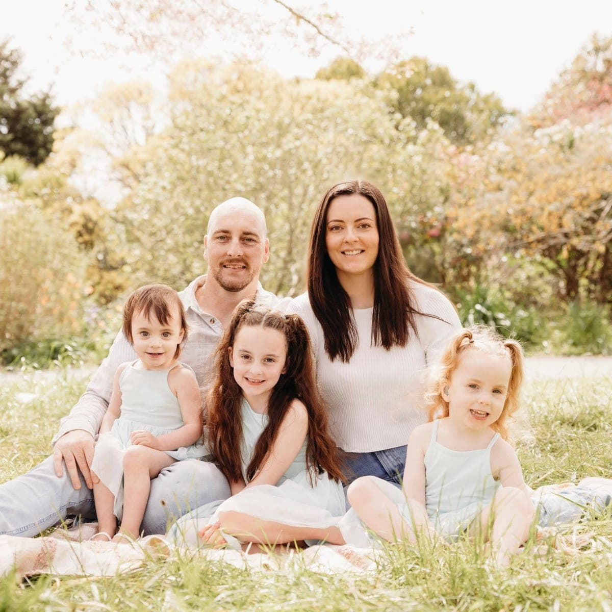 Joel with his wife and their three daughters, sitting on a picnic blanket on the grass in a garden