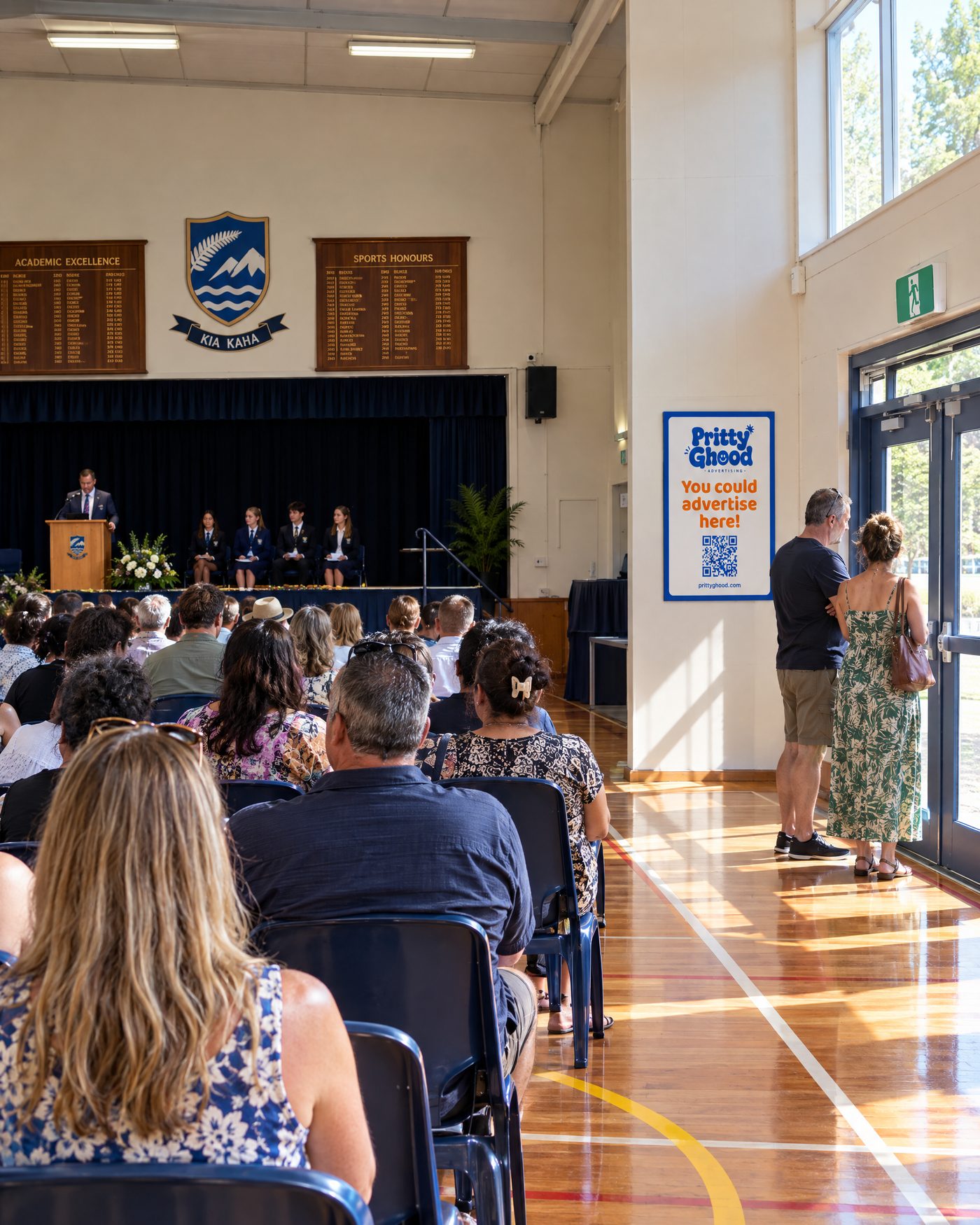 Families seated at a school prizegiving with a framed Pritty Ghood poster by the hall doors