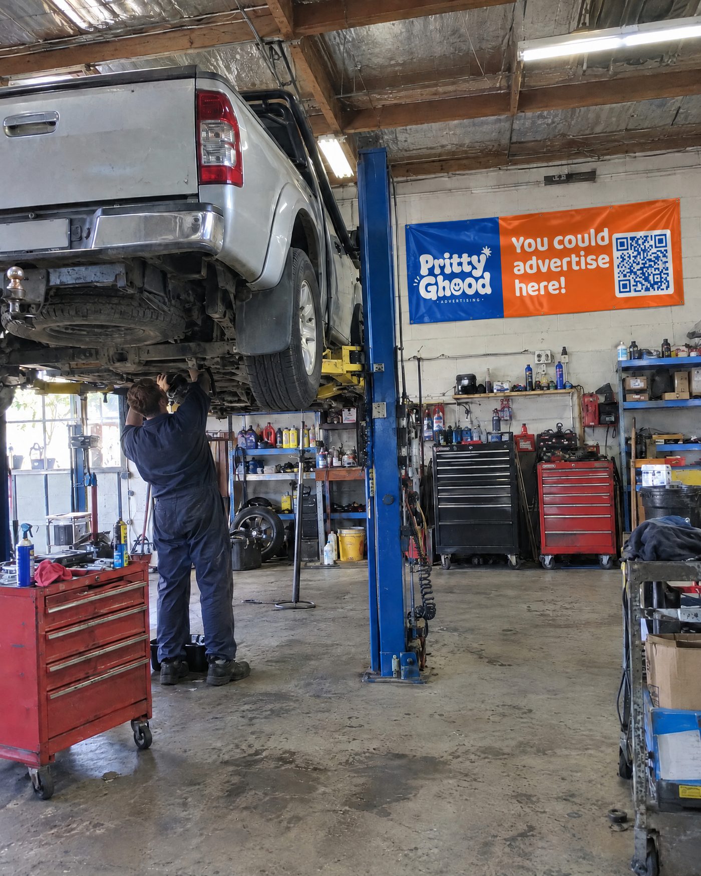 A mechanic working under a ute in a workshop with a Pritty Ghood banner on the wall